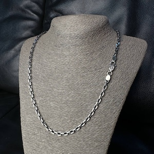 May include: A silver chain necklace with a box link design is displayed on a grey mannequin. The necklace is made of metal and has a clasp closure. The background is a dark leather surface.