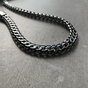 May include: A silver chain necklace with a woven design. The necklace is made of interlocking links, creating a thick, durable appearance. The chain is displayed on a grey surface.