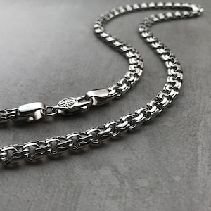 May include: A silver chain necklace with a box chain design. The chain is made of interlocking links that form a square pattern.