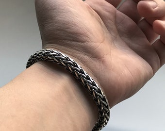 May include: A silver chain bracelet with a woven design. The bracelet is worn on a person's wrist.