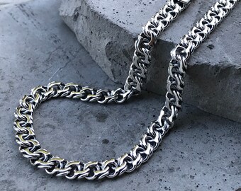 May include: A silver-colored chain necklace with a thick, textured design. The chain is made of interlocking links, creating a bold and eye-catching accessory. The necklace is displayed on a gray surface.