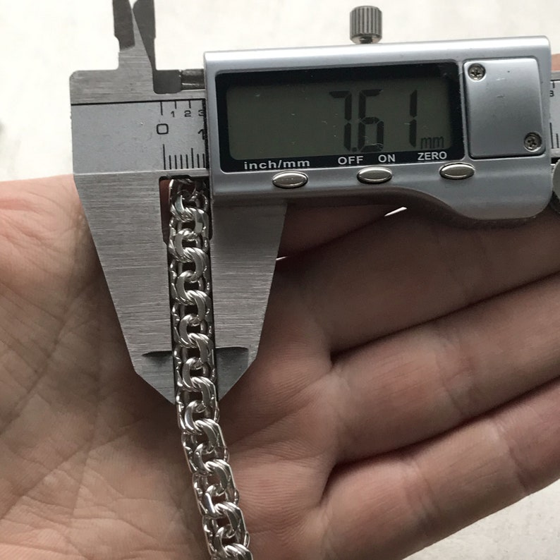 May include: A silver chain link bracelet being measured with a digital caliper. The caliper reads 7.61 mm.