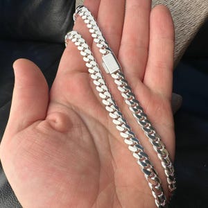 May include: Two silver curb chain necklaces, one thicker than the other, are laid out on a hand. The chains are made of interlocking links.
