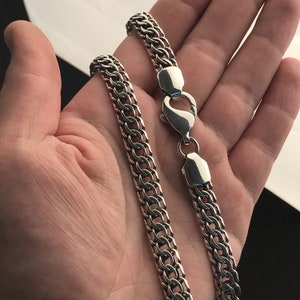 Peut inclure: Un collier en chaîne argent avec un fermoir à ressort. La chaîne est composée de deux rangées de maillons entrelacés.