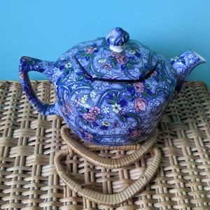 May include: A blue and pink floral teapot with a lid. The teapot is sitting on a woven wicker surface.