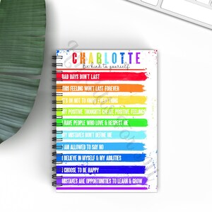 May include: A spiral bound notebook with a white cover and rainbow colored stripes. The notebook has the name "Charlotte" and the text "Be kind to yourself" printed on the cover. The notebook has affirmations printed on each stripe, such as "Bad days don't last" and "I choose to be happy".