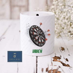 May include: White ceramic piggy bank with a black and white dartboard and red darts. The piggy bank has the name "ANDREW" written in green letters on the front.