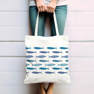 May include: A white canvas tote bag with a blue and white fish pattern. The bag has two handles and is being held by a person.