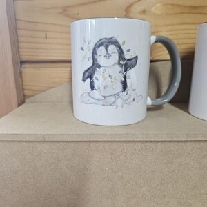 May include: White ceramic mug with a gray handle. The mug features a cartoon penguin wearing a string of lights. The penguin is standing on a snowy surface.