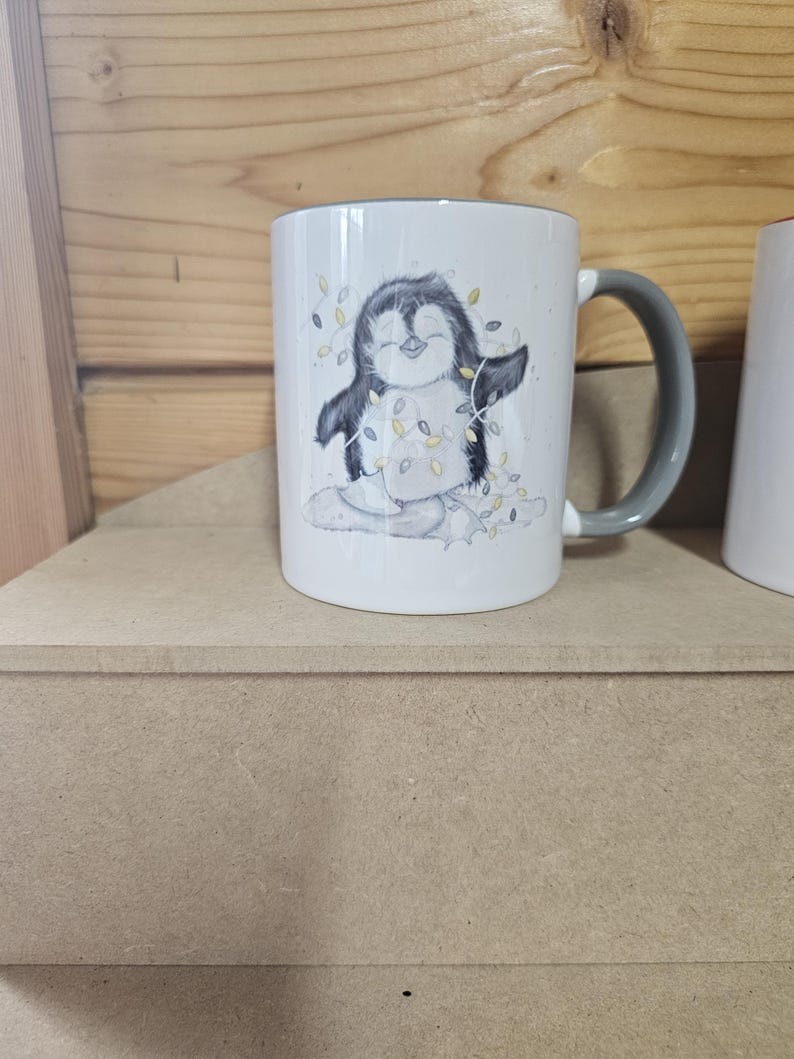 May include: White ceramic mug with a gray handle. The mug features a cartoon penguin wearing a string of lights. The penguin is standing on a snowy surface.