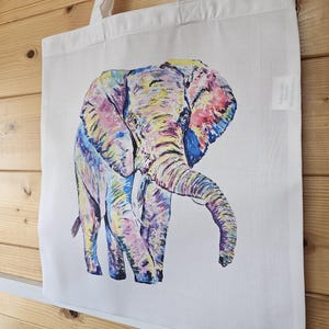 May include: A white tote bag with a colorful hand-painted illustration of an elephant. The elephant is painted in shades of pink, yellow, blue, and green.