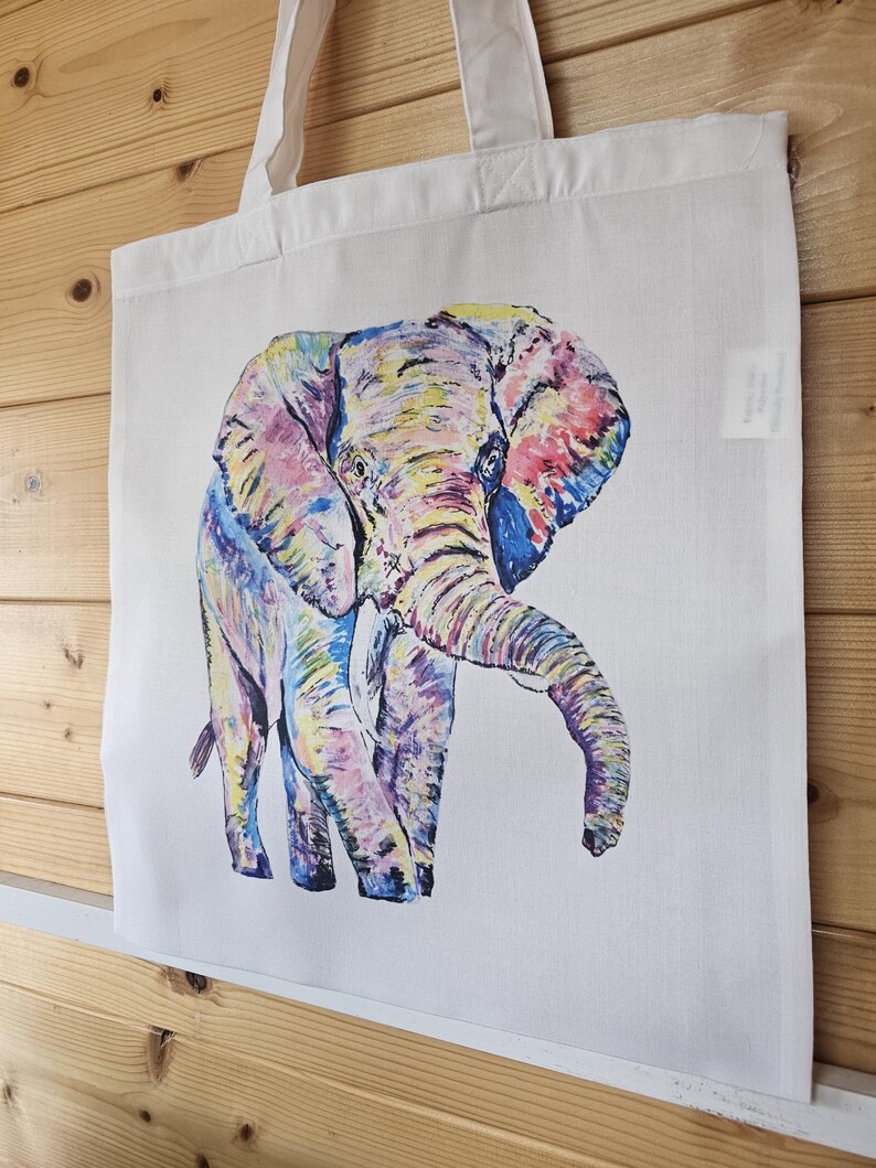 May include: A white tote bag with a colorful hand-painted illustration of an elephant. The elephant is painted in shades of pink, yellow, blue, and green.
