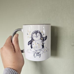 May include: White ceramic mug with a gray handle. The mug features a cartoon penguin wearing a string of lights and the name "Olly" printed below the penguin.