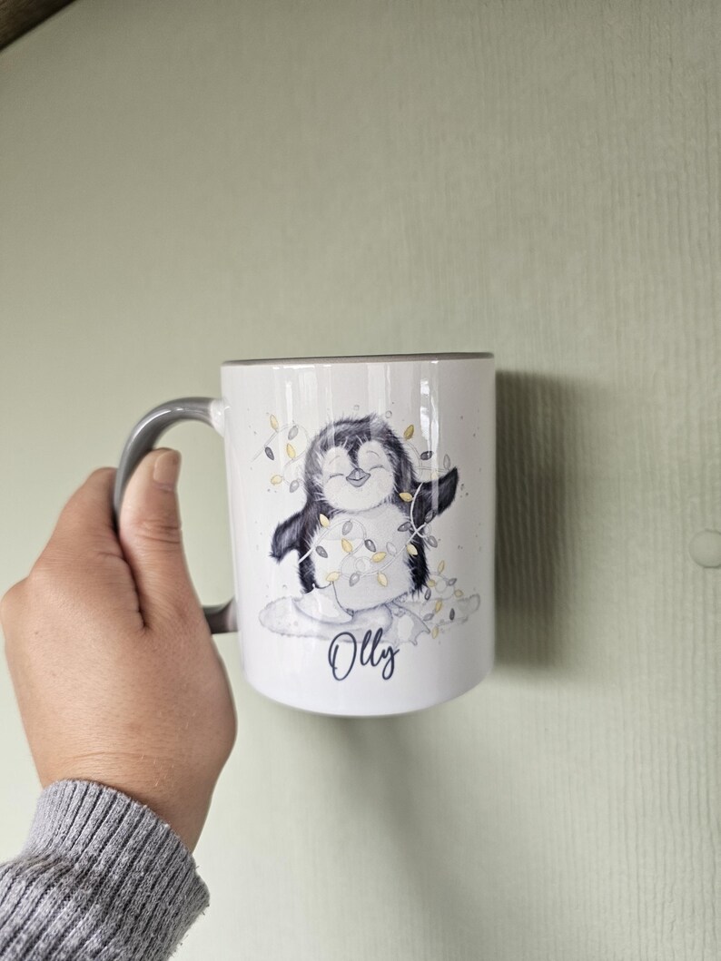 May include: White ceramic mug with a gray handle. The mug features a cartoon penguin wearing a string of lights and the name "Olly" printed below the penguin.