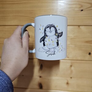 May include: White ceramic mug with a gray handle. The mug features a cartoon penguin wearing a string of lights and the name "Sarah" printed on the bottom.