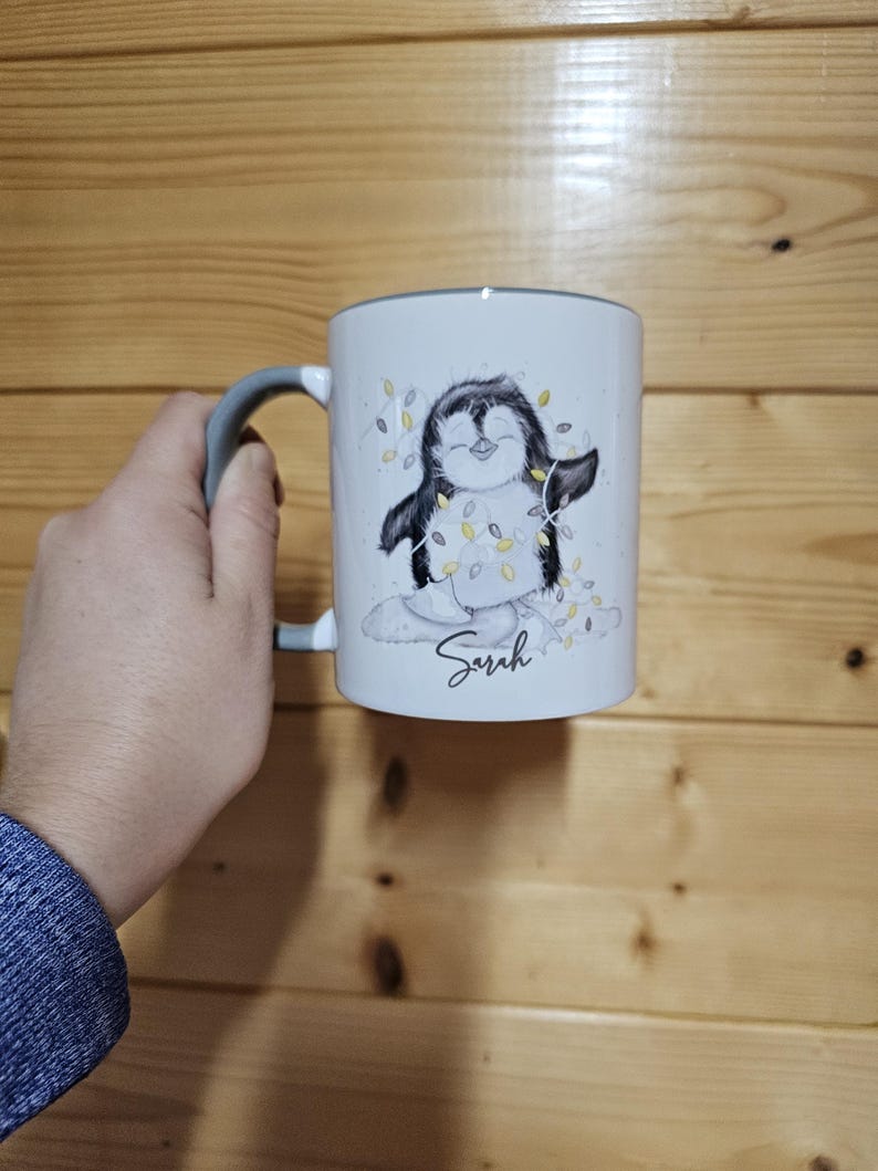 May include: White ceramic mug with a gray handle. The mug features a cartoon penguin wearing a string of lights and the name "Sarah" printed on the bottom.