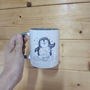 May include: A white ceramic mug with a gray rim and handle. The mug features a cartoon penguin wearing a string of lights.