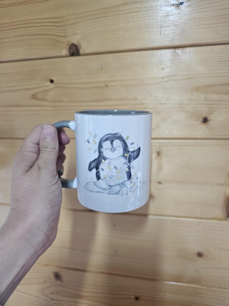 May include: A white ceramic mug with a gray rim and handle. The mug features a cartoon penguin wearing a string of lights.