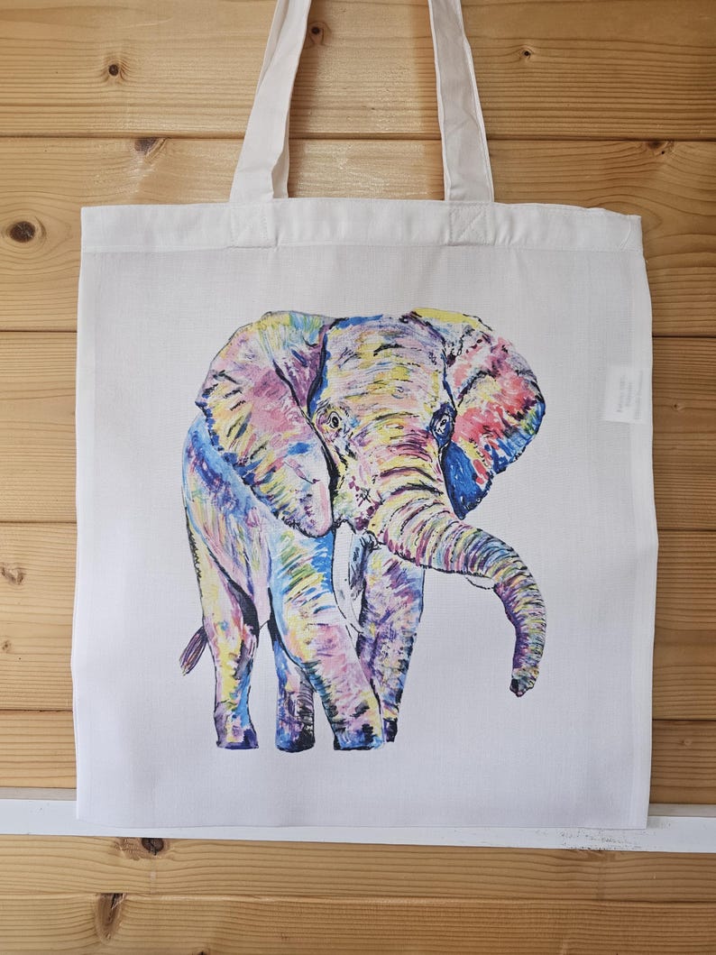 May include: A white tote bag with a colorful illustration of an elephant. The elephant is painted in shades of pink, yellow, blue, and purple.