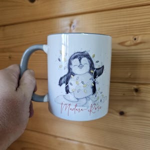 May include: White ceramic mug with a gray handle. The mug features a cartoon penguin wearing a string of Christmas lights. The text "Madara-Rose" is printed in red at the bottom of the mug.