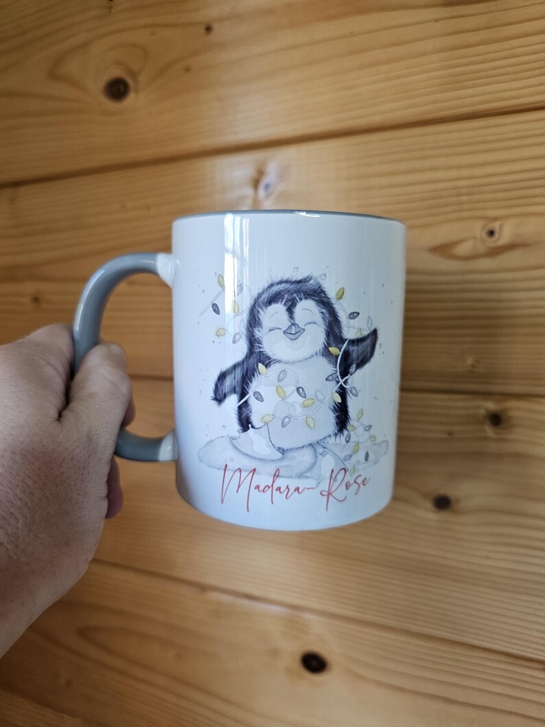May include: White ceramic mug with a gray handle. The mug features a cartoon penguin wearing a string of Christmas lights. The text "Madara-Rose" is printed in red at the bottom of the mug.