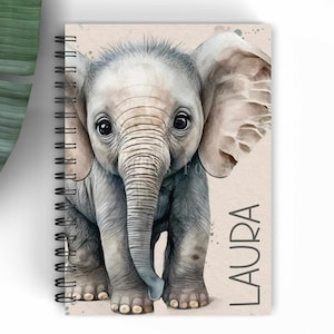 May include: A spiral bound notebook featuring a watercolour illustration of a baby elephant on the front cover. The elephant is grey with a light brown trunk and ears. The name "LAURA" is printed in grey text on the right side of the cover.