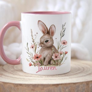 May include: A white ceramic mug with a pink rim and handle. The mug features a watercolour illustration of a brown bunny sitting in a field of pink flowers. The name "Lauren" is written in pink below the bunny.