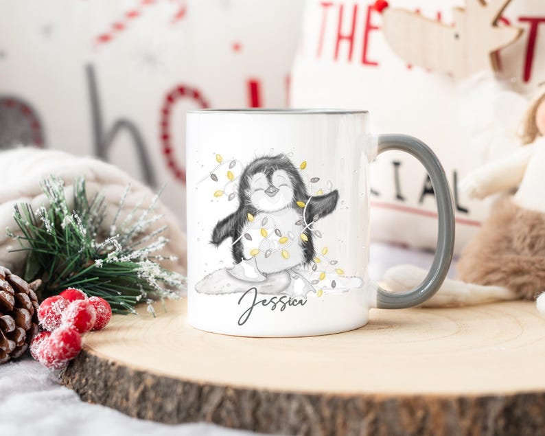 May include: White ceramic mug with a black and white illustration of a penguin wearing a string of lights. The mug has a black rim and handle and the name "Jessica" is written on the bottom.