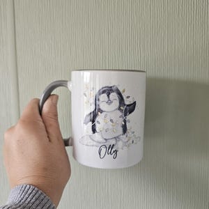 May include: White ceramic mug with a gray handle. The mug features a cartoon penguin wearing a string of lights and the name "Olly" written below.