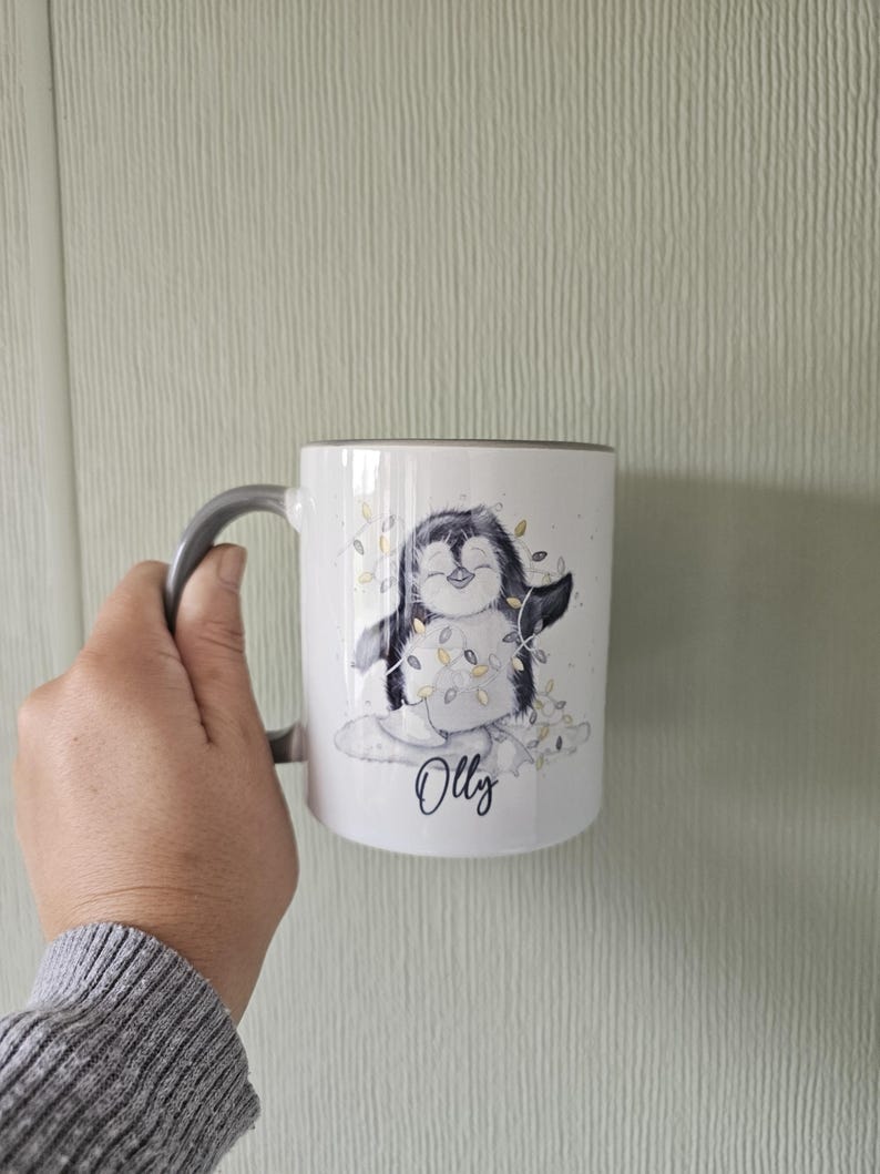 May include: White ceramic mug with a gray handle. The mug features a cartoon penguin wearing a string of lights and the name "Olly" written below.