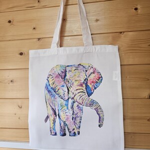 May include: A white canvas tote bag with a colorful illustration of an elephant. The elephant is painted in shades of pink, purple, blue, yellow, and green.