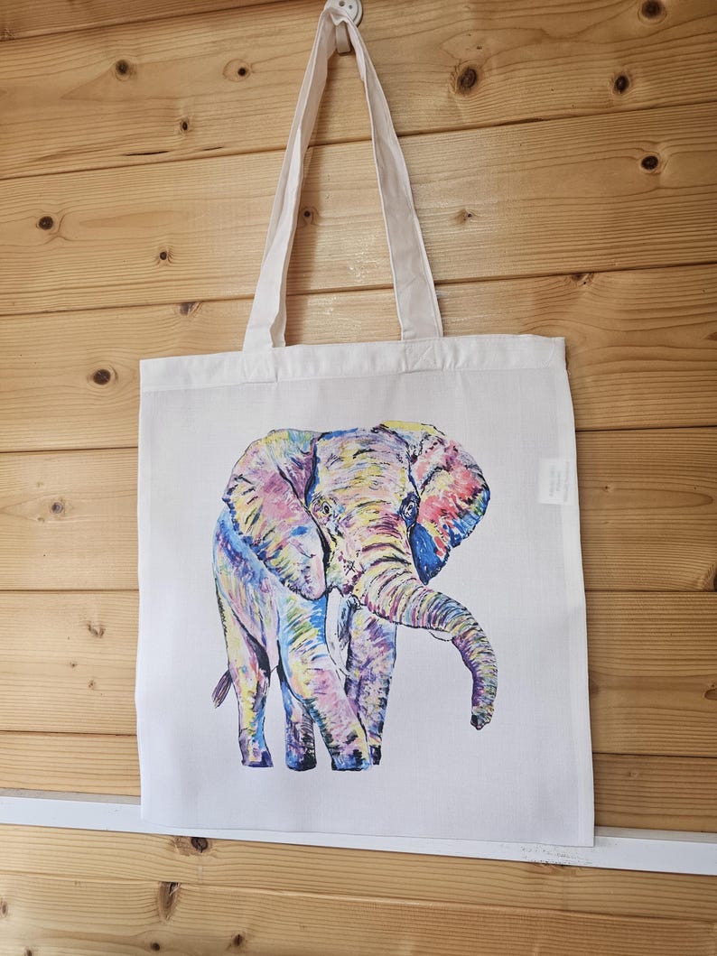 May include: A white canvas tote bag with a colorful illustration of an elephant. The elephant is painted in shades of pink, purple, blue, yellow, and green.
