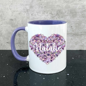 May include: White ceramic mug with a purple rim and handle. The mug features a heart-shaped design made of purple flowers with the name "Natalie" in white text.