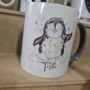 May include: White ceramic mug with a black and white illustration of a penguin holding a string of lights. The mug has a black handle and the name "Tisha" written in cursive on the bottom.