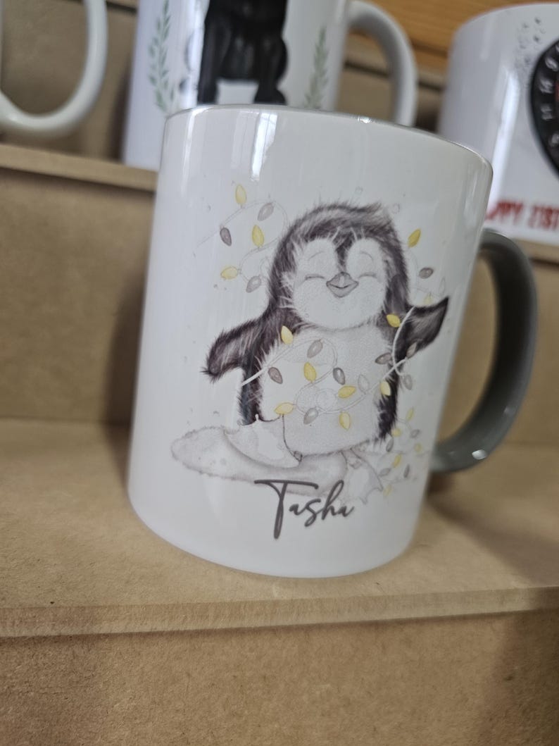 May include: White ceramic mug with a black and white illustration of a penguin holding a string of lights. The mug has a black handle and the name "Tisha" written in cursive on the bottom.