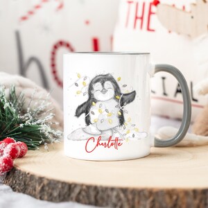May include: White ceramic mug with a gray handle. The mug features a black and white illustration of a penguin wearing a string of Christmas lights. The text "Charlotte" is printed in red below the penguin.