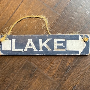 May include: A rectangular wooden sign with the word "LAKE" in white capital letters and a white arrow pointing right. The sign is painted navy blue with a distressed finish and has a jute rope for hanging.