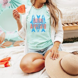Puede incluir: Camiseta verde claro con un gr&aacute;fico de tres peces azules y las palabras "BUON APPETITO" en rojo. La persona sostiene una rodaja de sand&iacute;a. Tambi&eacute;n se ven un sombrero de paja grande y un flotador floral.