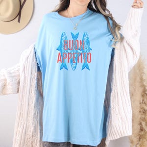 Puede incluir: Camiseta azul claro con tres peces azules estilizados y la frase roja "BUON APPETITO". Esta camiseta informal es ideal para el uso diario. El dise&ntilde;o est&aacute; centrado en la parte delantera de la camiseta.