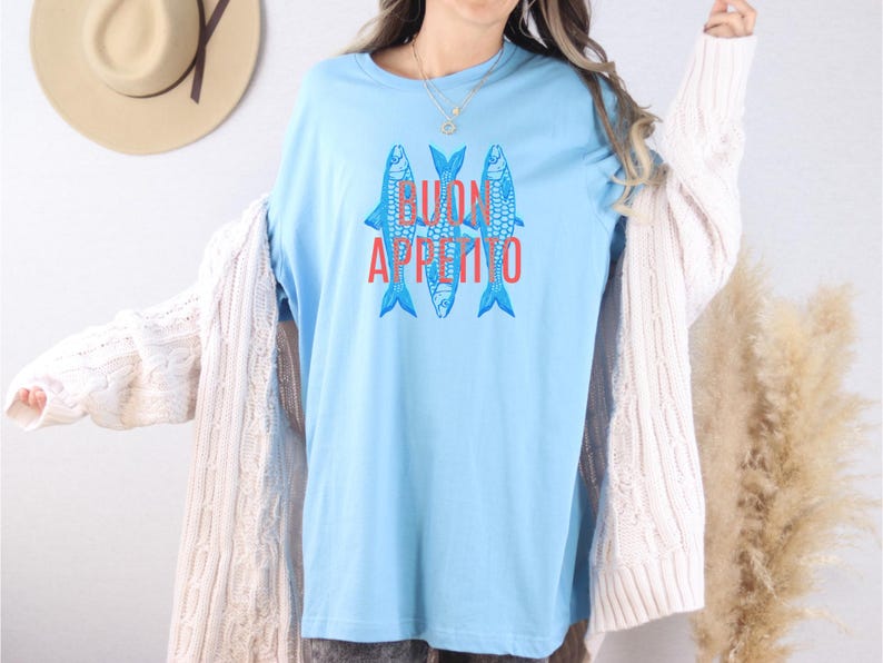 Puede incluir: Camiseta azul claro con tres peces azules estilizados y la frase roja "BUON APPETITO". Esta camiseta informal es ideal para el uso diario. El dise&ntilde;o est&aacute; centrado en la parte delantera de la camiseta.