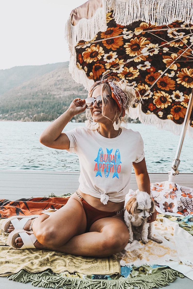 Puede incluir: Una mujer con gafas de sol y una camiseta blanca con un gr&aacute;fico de pescado y las palabras "Buon Appetito" est&aacute; sentada en un barco. Est&aacute; bajo una sombrilla floral. Un perro peque&ntilde;o est&aacute; sentado a su lado. La escena es al aire libre con un lago y monta&ntilde;as al fondo.