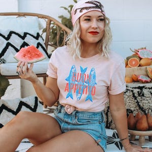 Puede incluir: Camiseta rosa claro con la inscripci&oacute;n "BUON APPETITO" y tres peces azules. La persona lleva un pa&ntilde;uelo rosa y pantalones cortos de mezclilla. Una rodaja de sand&iacute;a se sostiene en la mano. Frutas y cojines decorativos en el fondo.