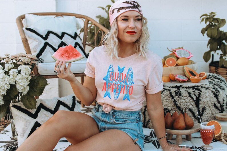 Puede incluir: Camiseta rosa claro con la inscripci&oacute;n "BUON APPETITO" y tres peces azules. La persona lleva un pa&ntilde;uelo rosa y pantalones cortos de mezclilla. Una rodaja de sand&iacute;a se sostiene en la mano. Frutas y cojines decorativos en el fondo.