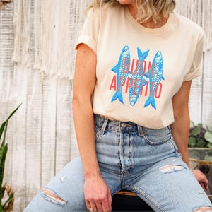 Puede incluir: Camiseta color crema con un gr&aacute;fico de tres peces azules y la frase "BUON APPETITO" en rojo. La camiseta se combina con vaqueros azules claros rotos. El estilo es informal y relajado.