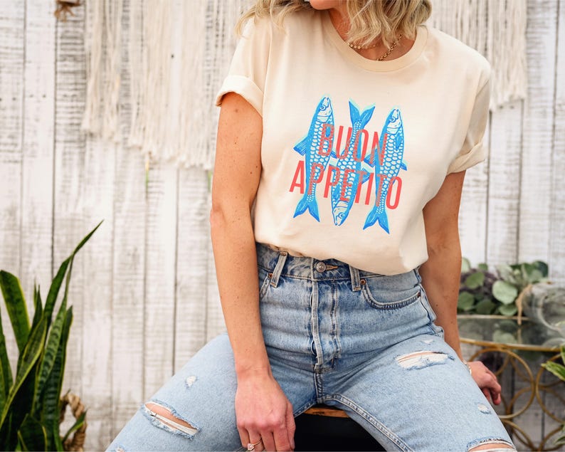 Puede incluir: Camiseta color crema con un gr&aacute;fico de tres peces azules y la frase "BUON APPETITO" en rojo. La camiseta se combina con vaqueros azules claros rotos. El estilo es informal y relajado.