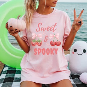 May include: A light pink t-shirt with the words "Sweet & Spooky" in pink script. The shirt features a graphic of cherries and strawberries with bows. A pink pumpkin and a white ghost plushie are also visible.