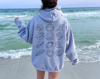 May include: A pale grey hoodie featuring black seashell designs on the back. The hoodie has a hood and long sleeves. The person is standing on a sandy beach with the sea in the background.