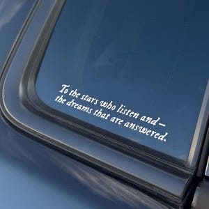 Op de afbeelding: Witte sticker op een autoraam met de tekst: "To the stars who listen and - the dreams that are answered." De sticker is in een scriptlettertype en is geplaatst op een donkerblauwe autoraam.