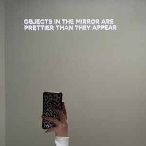 Può includere: Un muro bianco con il testo "OBJECTS IN THE MIRROR ARE PRETTIER THAN THEY APPEAR" in lettere bianche. Una mano tiene una custodia per telefono nera con un motivo leopardato.