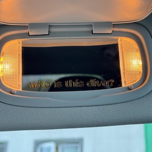 Pode incluir: Um espelho de viseira de carro com o texto dourado "who is this diva?". O espelho é emoldurado por uma luz amarela quente. A viseira é de cor cinza claro.